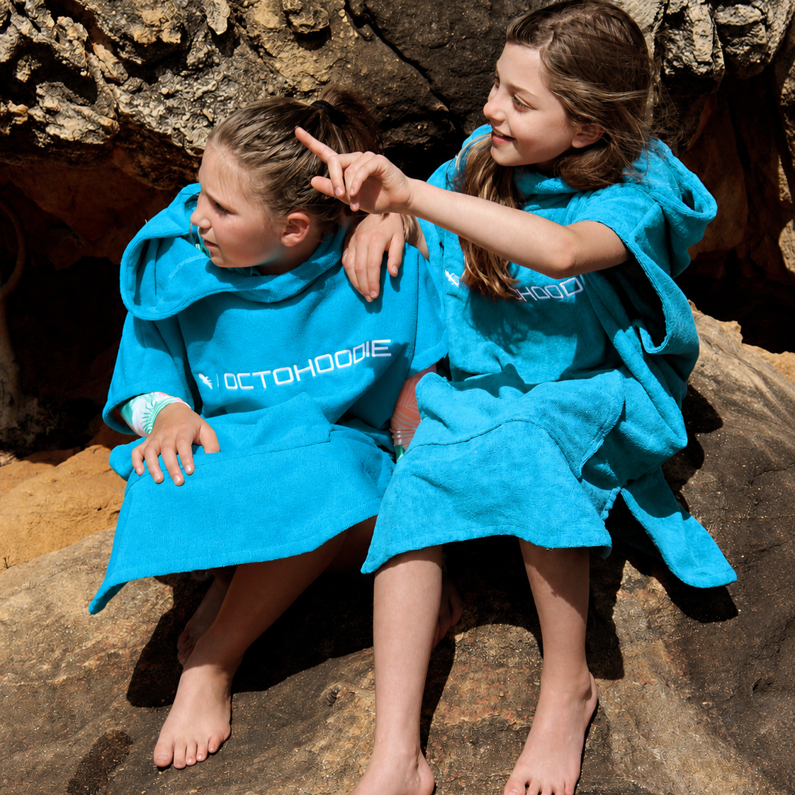 Two children sitting barefoot on a rocky outdoor surface, wearing bright turquoise Octohoodies