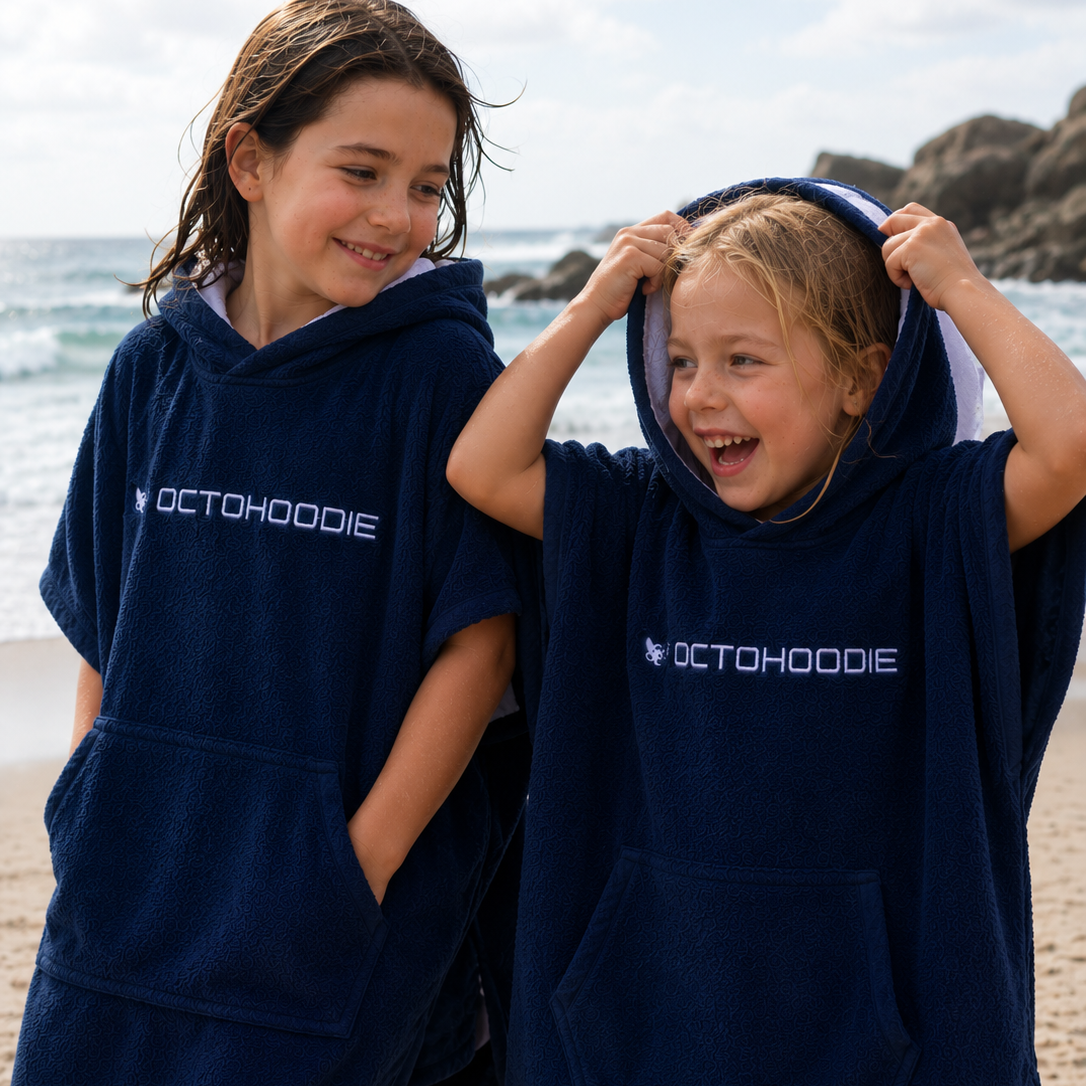 Two children wearing navy blue Octohoodie ponchos on a beach.