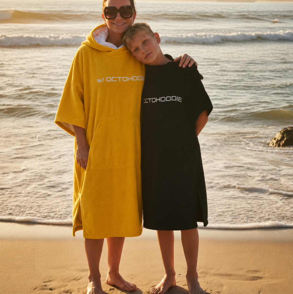 An adult and a child standing on a sandy beach at sunset, each wearing Octohoodies