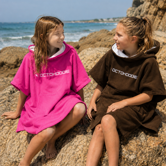 Two girls sitting on large rocks by the ocean, wearing short-sleeve Octohoodies