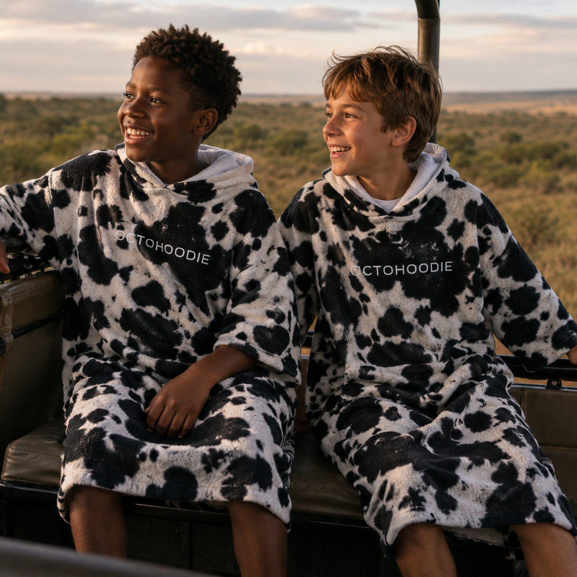 Two children wearing Nguni patterned polar fleece winter hoodies sitting in a vehicle with a safari landscape in the background.