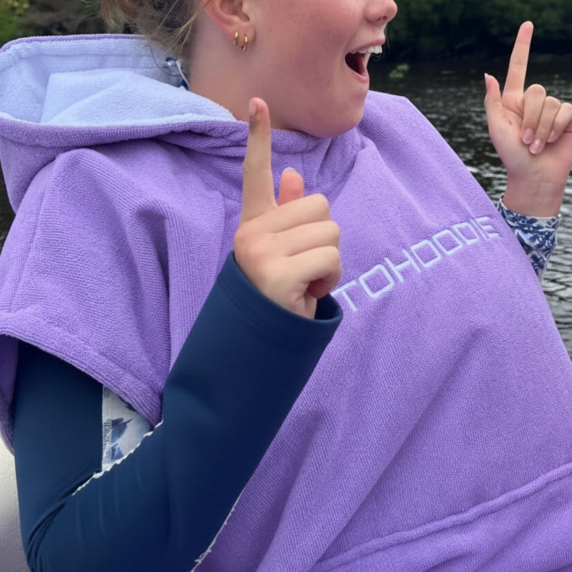 A person outdoors wrapped in a lavender-purple short sleeve Octohoodie swimming poncho. The background shows water and green foliage.