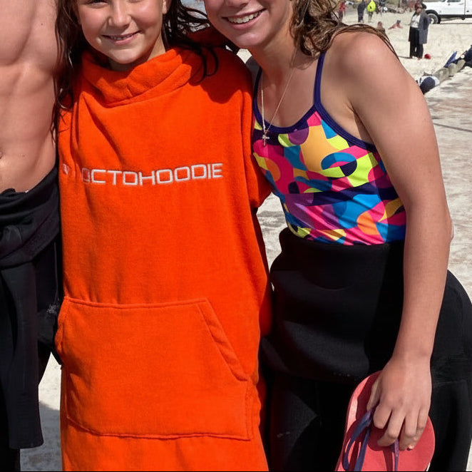 Three people standing on a beach boardwalk wearing swimwear and wetsuits, with a bright orange Octohoodie